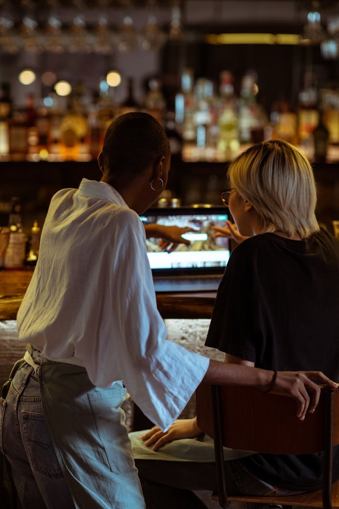 Two women browsing a laptop together at a cozy café bar, seen from behind in a relaxed atmosphere.